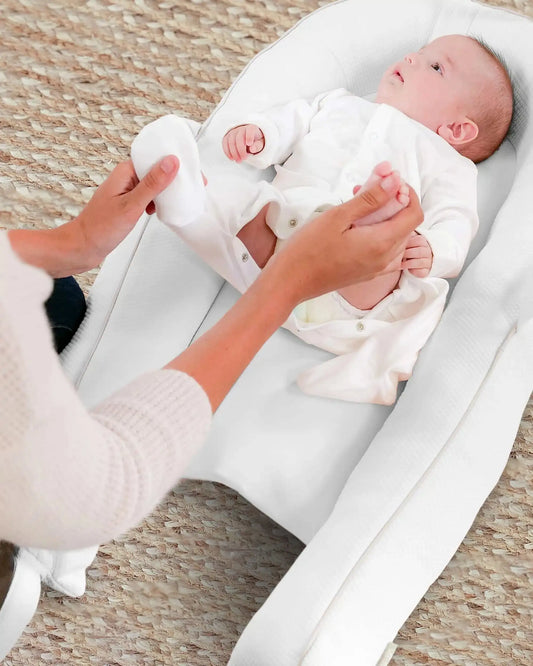 Baby lying in ergonomic white pillow nest as adult changes nappy on woven rug