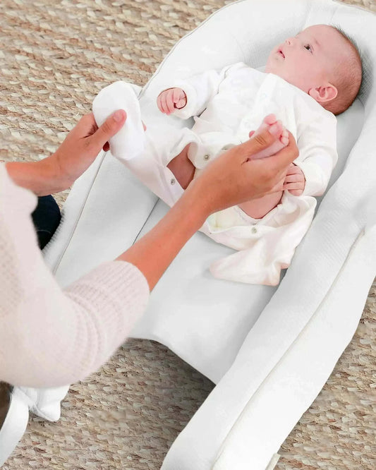 Baby lying in ergonomic white pillow nest as adult changes nappy on woven rug