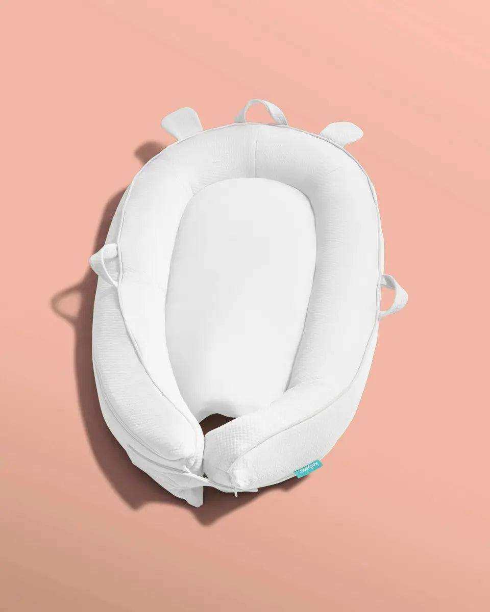 White ergonomic baby sleep nest pillow with handles on a peach background